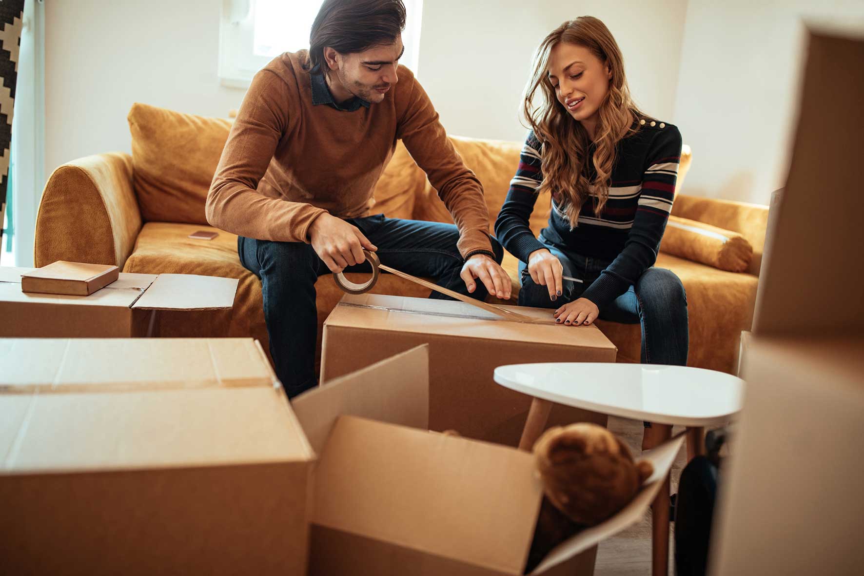 Two people sit on a tan sofa packing a cardboard box, using tape and scissors in a cozy living room.