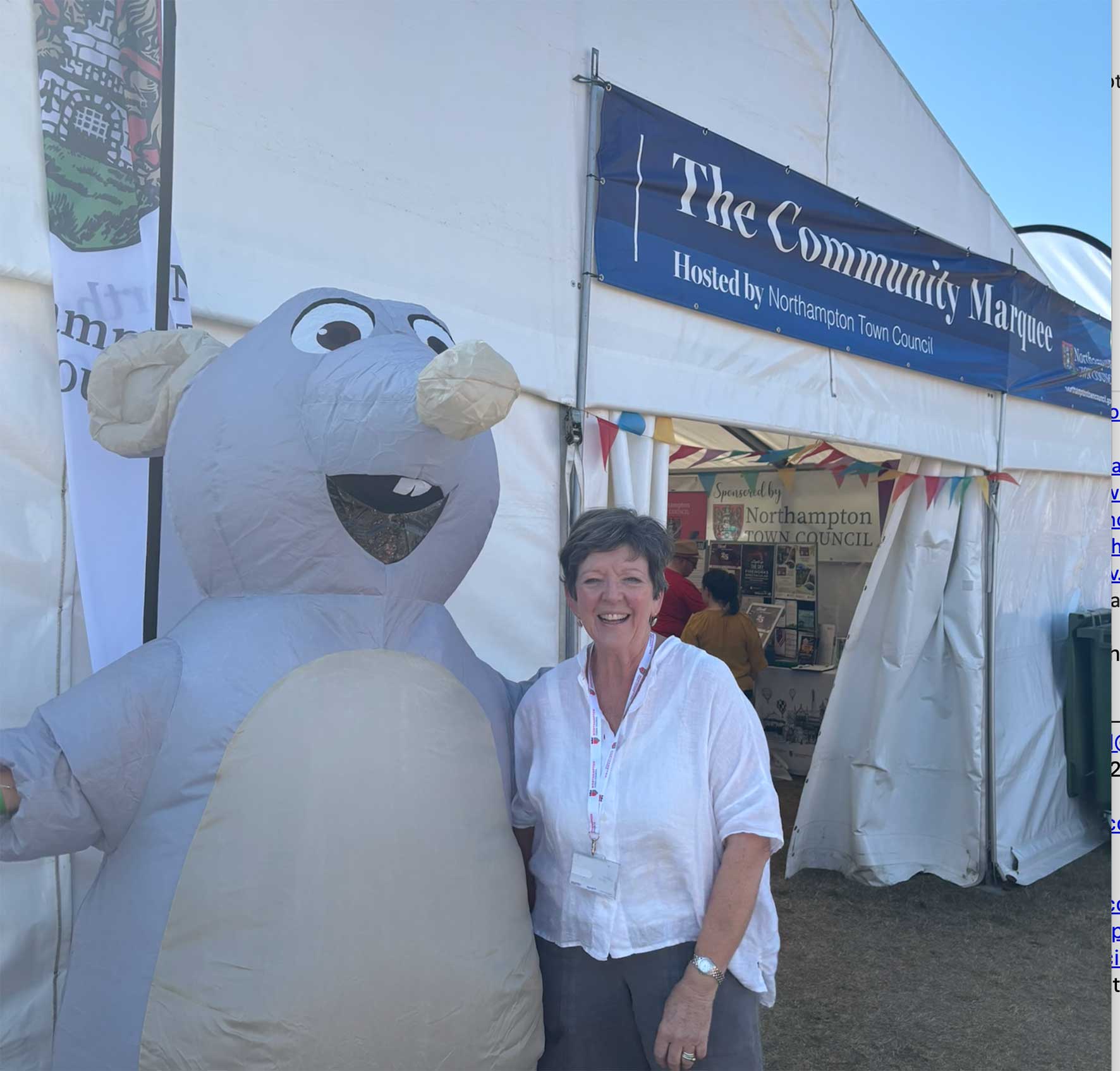 Smiling woman in a white blouse stands next to a large inflatable gray snowman at a community marquee tent, with a blue banner that reads 'The Community Marquee'.