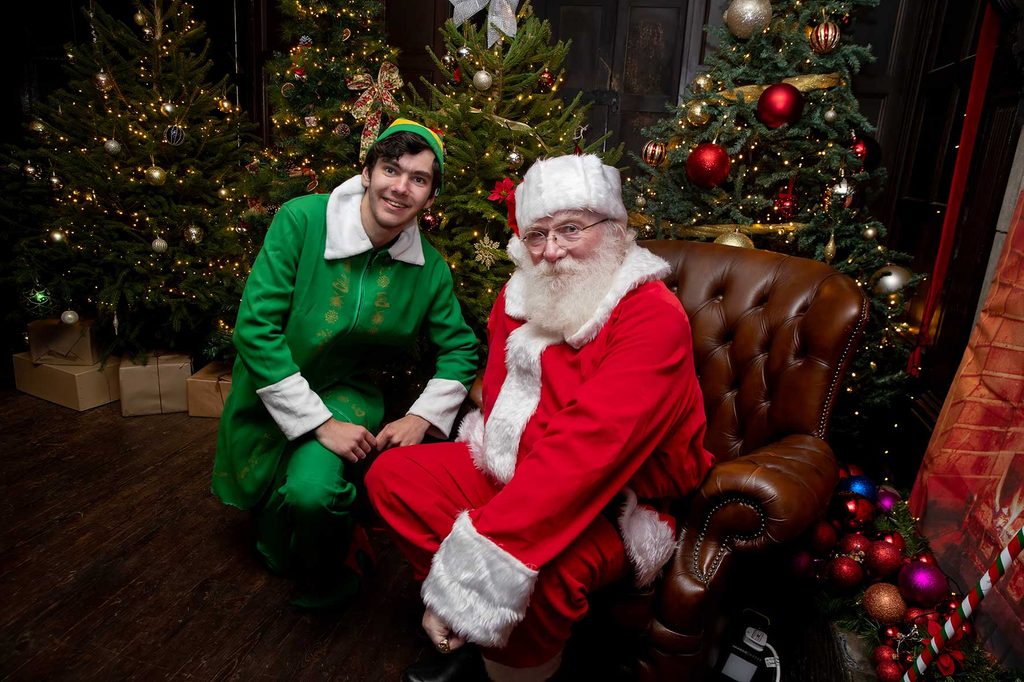 Delapre-Christmas-Market--2 | NN Pulse Smiling Santa in a red suit sitting on a brown leather sofa next to a cheerful elf in green, with Christmas trees, lights, and wrapped presents in the background.